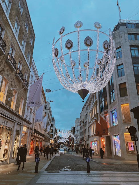 A city street during the evening with tall, modern buildings on either side, featuring luxury retail storefronts such as Dior and Louis Vuitton. The street is decorated with elaborate, illuminated chandeliers hanging overhead, made of multiple rings and teardrop-shaped elements, creating a festive atmosphere. Pedestrians are walking along the sidewalk, some in groups and others alone, amidst seasonal decorations and bright shop displays. The wide, clean pavement is marked with a bicycle lane, and the scene is lit by natural twilight and decorative lighting, highlighting the crispness and cleanliness of the urban environment. This scene exemplifies refined surface cleaning and maintenance practices typical of high-end shopping districts, where Mayfair Carpet Cleaning’s expertise in deep cleaning and hygiene supports the pristine appearance of commercial spaces in central London during special occasions or festive seasons.