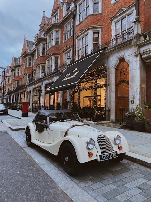 A classic vintage car, painted in cream with black accents, is parked on a city street adjacent to a row of red-brick townhouses with white-framed bay windows and ornate stone details. The car features a rounded front grille and large circular headlights, with a black soft top in the closed position. The street is paved with grey cobblestones and concrete, with a narrow sidewalk lined by potted plants outside a boutique shop that has a black awning displaying the Mayfair Carpet Cleaning logo, highlighting the area's elegant urban setting. The scene is illuminated by natural daylight, emphasizing the clean and well-maintained appearance of the surroundings, reflecting the high standards of surface cleaning and maintenance promoted by Mayfair Carpet Cleaning.