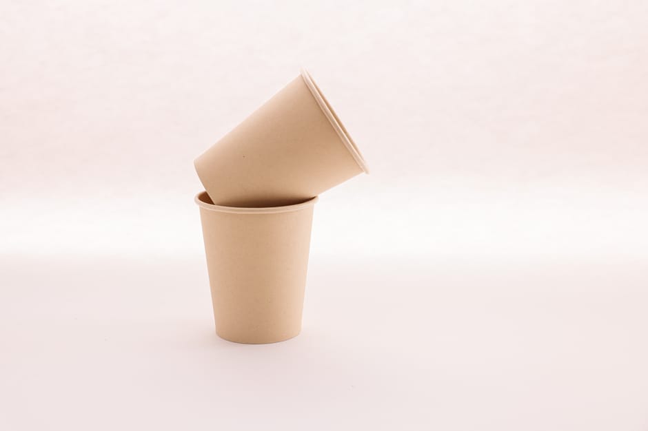 A cylindrical metal mesh waste bin filled with crumpled white paper is positioned on a beige carpeted floor within a room. A few crumpled sheets of paper are scattered on the carpet nearby. The backgr