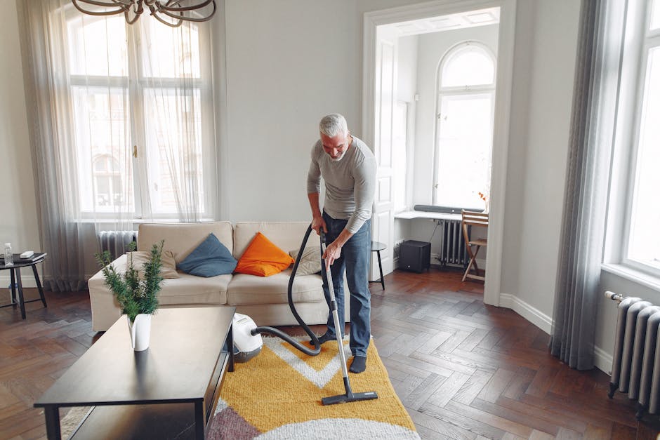 A professional cleaner dressed in full white protective suit, face mask, and blue gloves performing surface cleaning with a steam or fogger device on a plush beige carpet in a modern, well-lit living