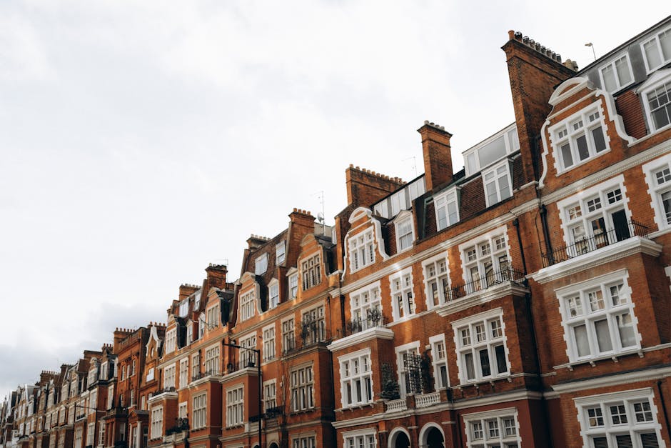 A red sports car parked on a city street in Mayfair, surrounded by mature buildings with brick and stone facades. The surface of the car is smooth and shiny, reflecting the nearby streetlights and the