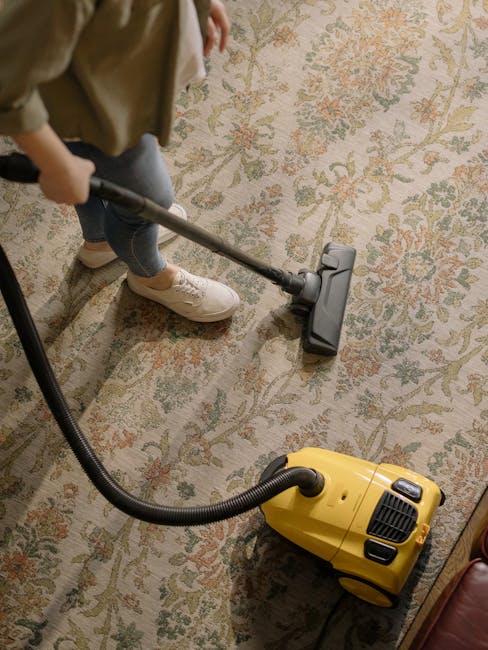 A person operating a yellow vacuum cleaner on a patterned, beige and green carpet in a room with natural lighting. The individual is wearing beige shoes, blue jeans, and a beige jacket, and is holding the vacuum's hose attached to a cleaning head that is actively being used on the carpet. The setting appears to be a residential space, with the vacuum cleaner and hose positioned at an angle that indicates active surface cleaning, typical of domestic maintenance by Mayfair Carpet Cleaning as part of their period home care at Mount Street flats. The scene emphasizes thorough surface cleaning to remove dust and dirt, maintaining the carpet's appearance and hygiene.
