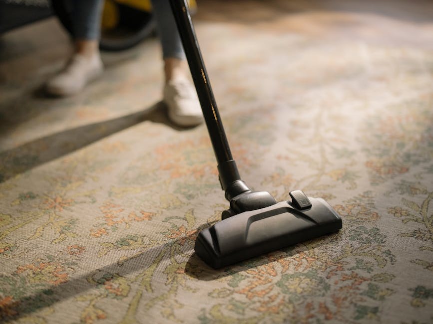Close-up of a vacuum cleaner head actively cleaning a traditional patterned rug with intricate floral designs in beige, green, and orange tones. The vacuum is positioned on a wooden floor section adjacent to the rug, with visible shadows indicating bright lighting. In the background, a person's legs and white shoes are partially visible, suggesting a domestic setting. The image highlights surface cleaning and maintenance of period home flooring, aligning with Mayfair Carpet Cleaning’s specialist services for historic properties in Mayfair.