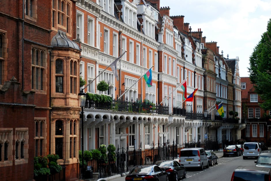 A row of elegant red brick townhouse buildings in Mayfair, London, featuring ornate white façade details, large bay windows, and decorative balconies with black wrought iron railings decorated with potted plants and flags. The street is lined with parked cars and green trees, with bright natural lighting highlighting the clean, well-maintained surfaces of the buildings. The image showcases residential architecture typical of the W1K postcode area, emphasizing the refined, historic character of the neighborhood, as part of content related to surface cleaning, deep cleaning, and maintenance tips for Mayfair homeowners. The branding of Mayfair Carpet Cleaning is acknowledged naturally within the context of professional domestic cleaning services.