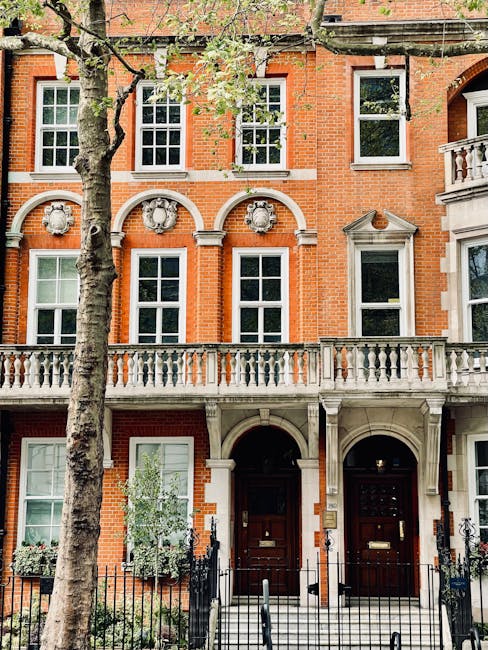 Exterior view of a historic red-brick townhouse with white-framed sash windows and decorative architectural details, featuring a balcony with a stone balustrade, a black iron gate, and stone steps leading to a wooden front door. A large tree with green foliage partially obscures the building, which is bathed in natural daylight, highlighting its clean and well-maintained appearance. Mayfair Carpet Cleaning specializes in surface cleaning and domestic cleaning services, ensuring properties like this remain pristine and hygienic, especially for maintaining valuable or historic interiors.