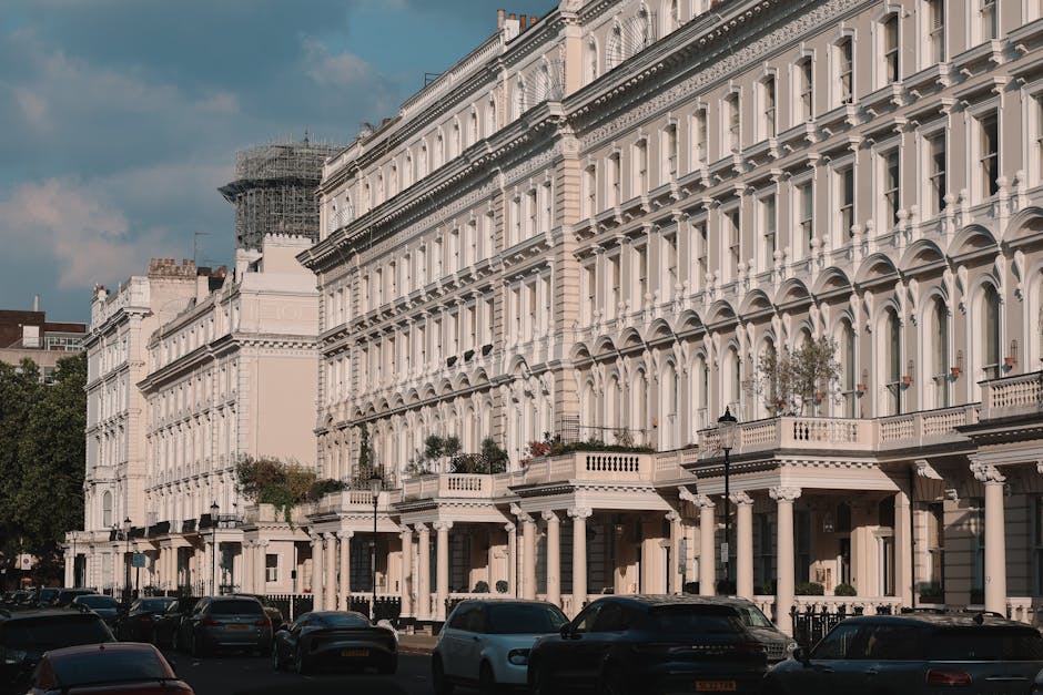 A row of elegant red brick townhouse buildings in Mayfair, London, featuring ornate white façade details, large bay windows, and decorative balconies with black wrought iron railings decorated with potted plants and flags. The street is lined with parked cars and green trees, with bright natural lighting highlighting the clean, well-maintained surfaces of the buildings. The image showcases residential architecture typical of the W1K postcode area, emphasizing the refined, historic character of the neighborhood, as part of content related to surface cleaning, deep cleaning, and maintenance tips for Mayfair homeowners. The branding of Mayfair Carpet Cleaning is acknowledged naturally within the context of professional domestic cleaning services.